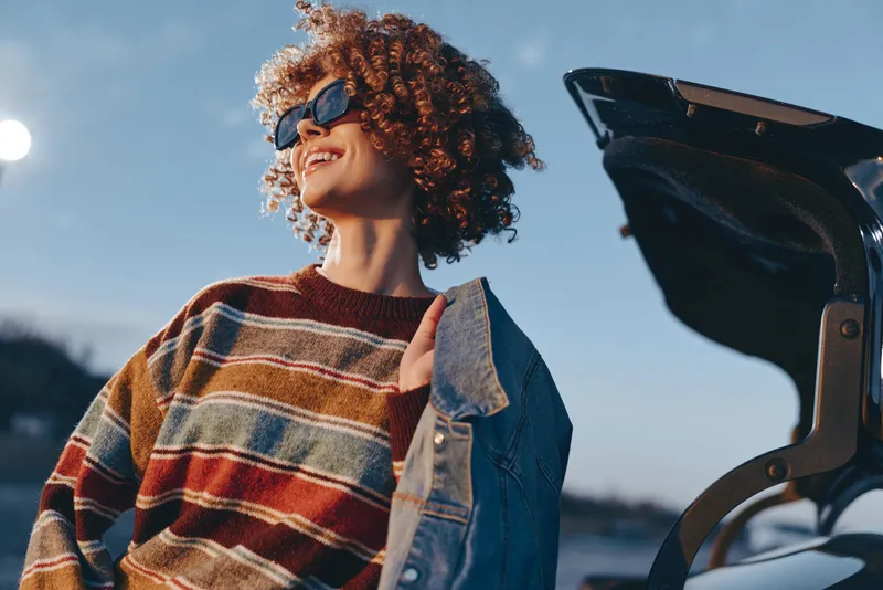 Woman lifestyle outdoors, curly hair, denim jacket, striped sweater, smile in sunlight, standing near beach horizon, enjoying seaside breeze, relaxed candid portrait with bright sky
