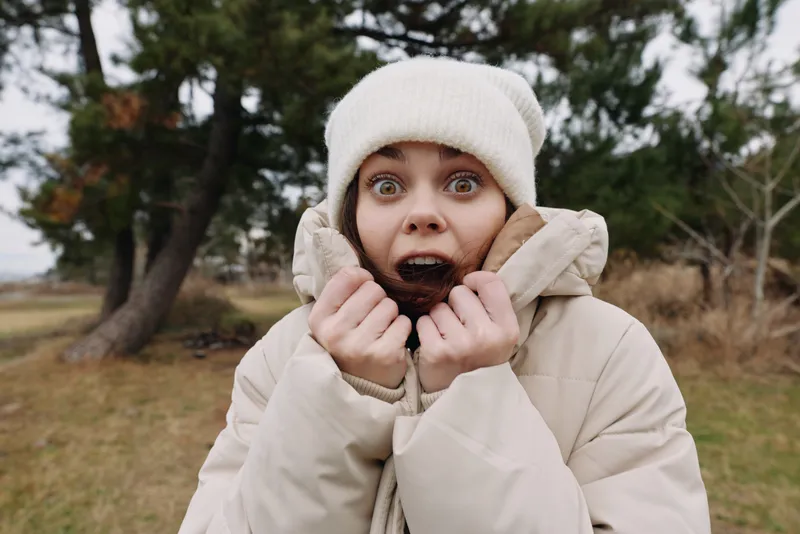 Woman outdoors in a winter coat and beanie with a surprised expression, gripping the collar, in a cold weather scene with bare trees and an open field on a chilly afternoon