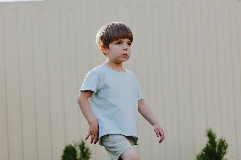 Young boy walking outdoors in a casual outfit, exuding curiosity and confidence in a natural setting surrounded by greenery.