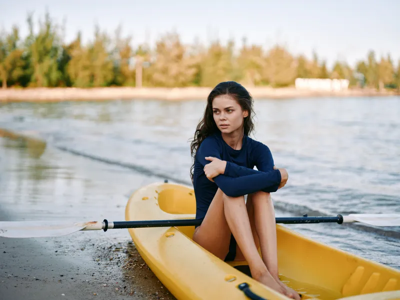 Young woman sits in a yellow kayak by the shore, arms wrapped around knees, gazing across calm water. Soft light, tranquil mood, introspective moment, coastal serenity and quiet reflection.
