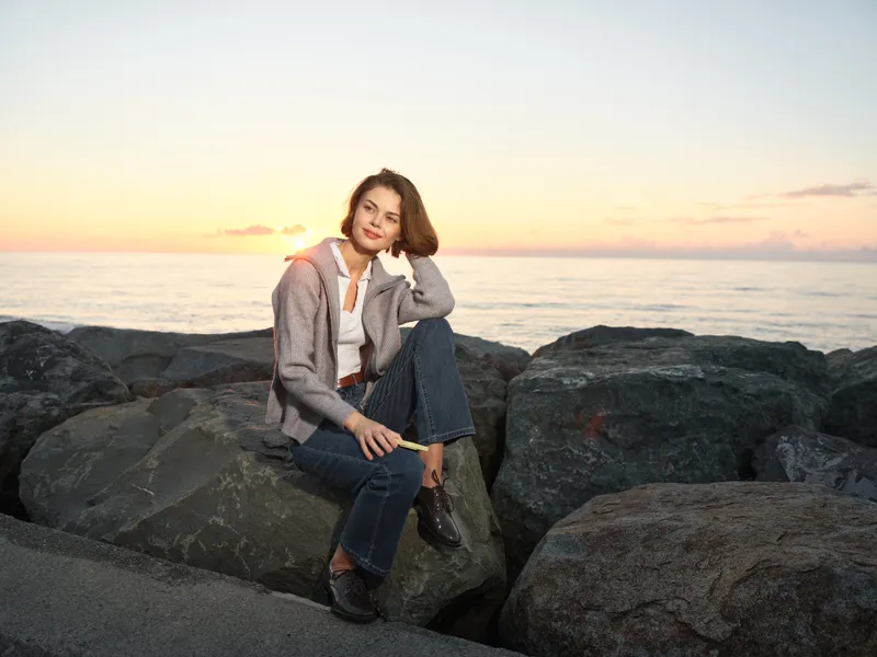 A woman sits on a rocky shoreline at sunset, wearing a casual jacket and jeans. She smiles softly, enjoying a peaceful moment by the sea, amid warm light and tranquil atmosphere.