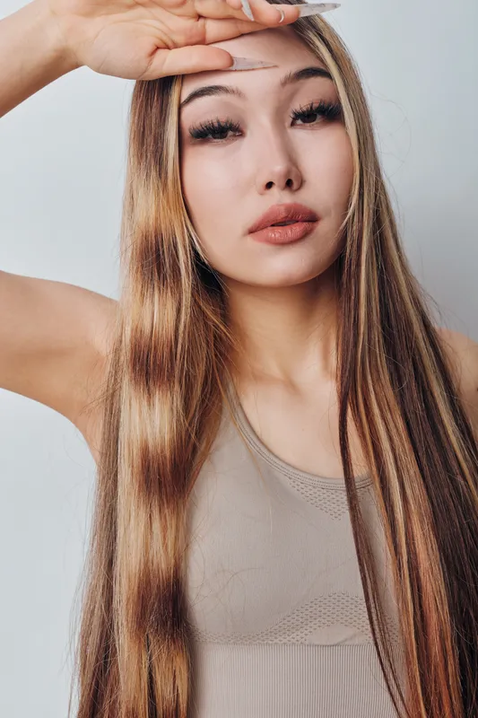 Young woman with long straight hair wearing beige tank top posing with hand shading eyes against light background. Female beauty, fashion, casual style and confident expression closeup.
