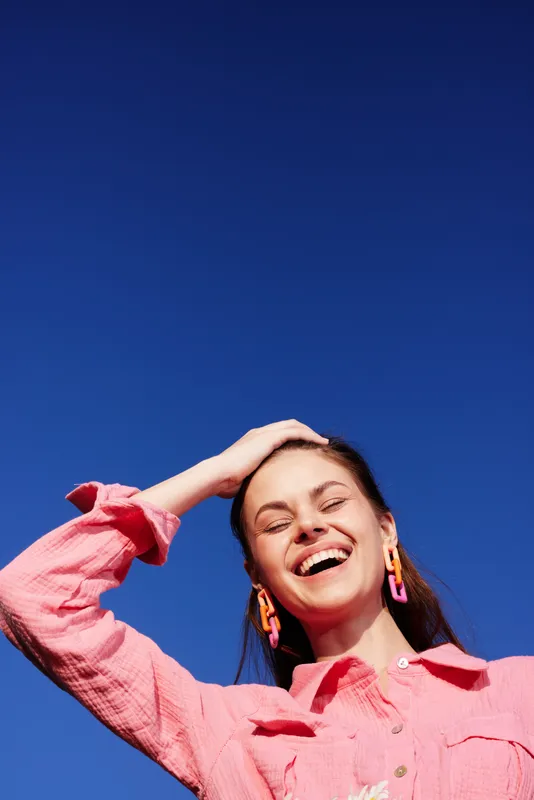 Joyful young woman in a pink shirt smiling against a clear blue sky, showcasing happiness and carefree emotions, ideal for positive lifestyle concepts