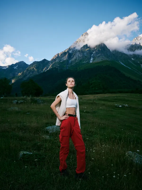 Hiker woman stands in a wide alpine meadow, red pants and white vest, hands on hips, looking toward distant peaks under a clear sky, encapsulating outdoor adventure and rugged landscape.
