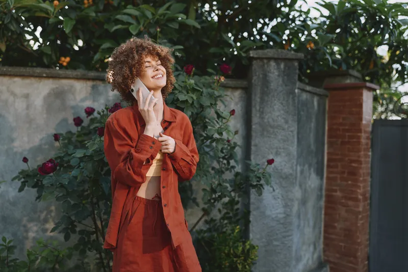 Young woman with curly hair stands outdoors in a rust blazer and pants, eyes closed, smiling as she touches her cheek in warm sunlight near a garden wall with blooming flowers, conveying a confident