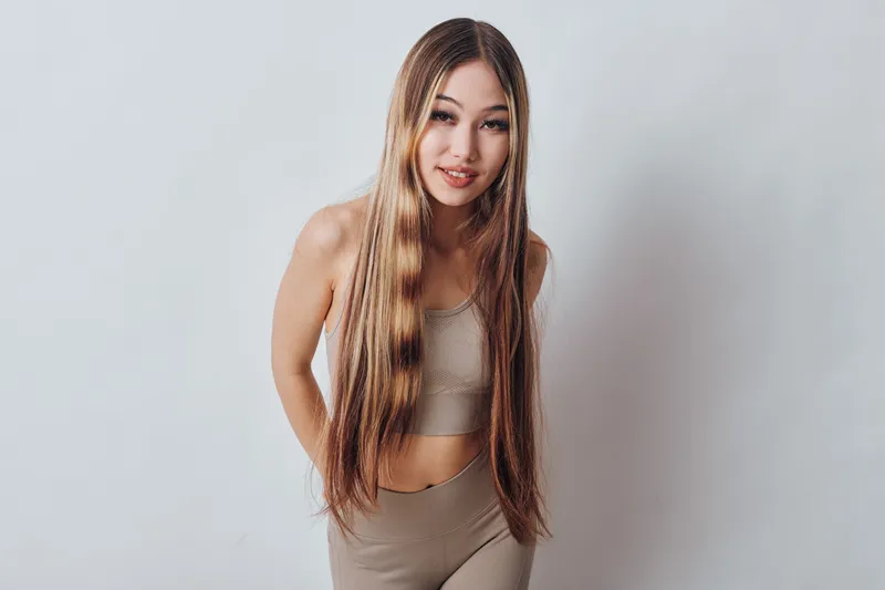Young woman with long straight hair wearing beige sporty outfit posing smiling looking at camera on plain light background in studio.