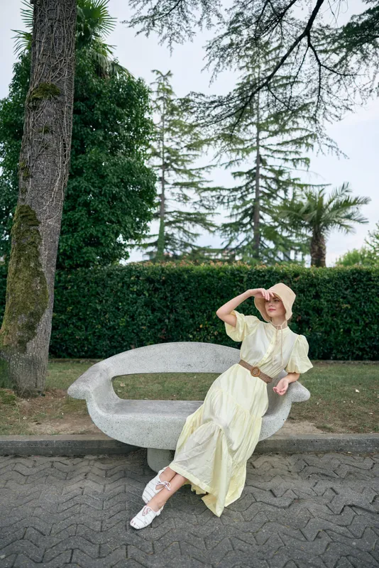 Woman on bench in park wearing flowing dress and sun hat, relaxation pose with natural light, authenticity and inclusivity in serene garden setting