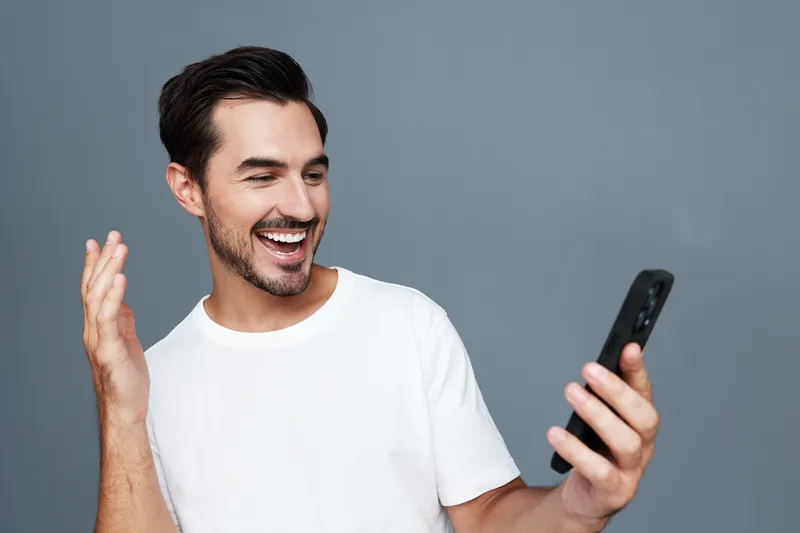 Cheerful smiling man in a casual white t-shirt looking at his smartphone with excitement, isolated on a solid gray background in a studio portrait. Happy lifestyle concept.