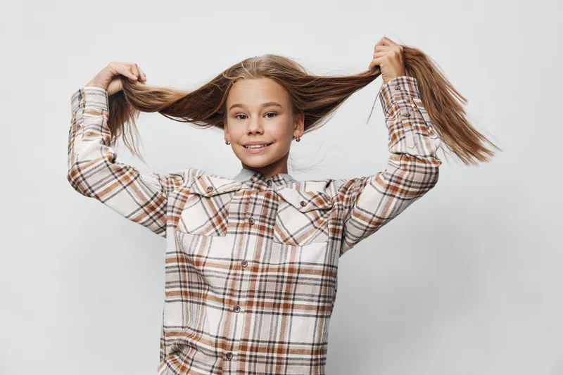 Girl child playing with long hair and smiling with plaid shirt on white background. Portrait of happy kid female with playful gesture and casual clothing for childhood fun.