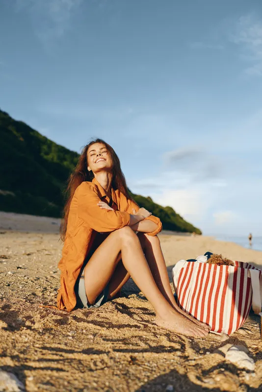 Beach woman sitting on sand in orange coat near seaside horizon, travel vacation mood, striped bag on ground beside her, warm sunlight, calm smile, outdoor leisure scene