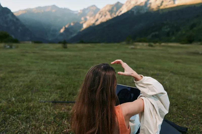 A person sits on a grassy field with a laptop, surrounded by mountains, enjoying outdoor work. Bright daylight and a calm atmosphere convey focus and creativity.