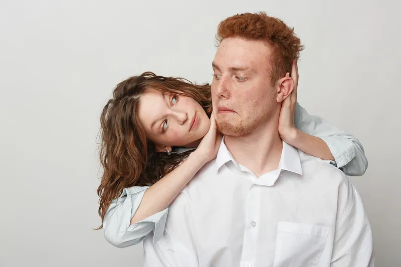 Curly woman embraces redhead man from behind, both wearing white shirts. The man looks surprised while the woman smiles gently. Concept of affection, relationship, and playful moment.