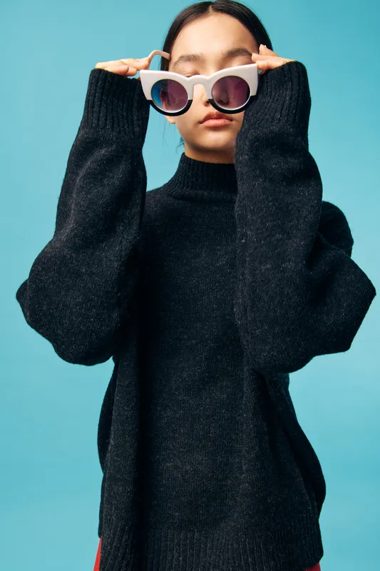 Woman with long hair wearing black oversized sweater holding white sunglasses on face against blue background, casual fashion style and confident pose in studio portrait.