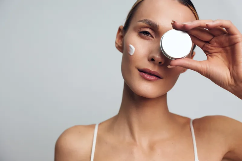 A young woman with clear skin holds a round container of cream near her face while applying moisturizer on her cheek. The background is plain and neutral, emphasizing skincare and beauty.