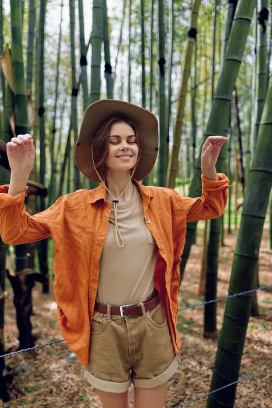 Woman in hat smiling among tall bamboo forest, enjoying outdoors with orange jacket, casual shorts and relaxed travel vibe. Portrait, nature walk and peaceful adventure.
