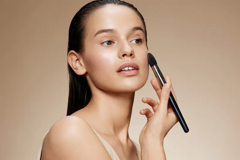 Young woman applying makeup with a brush, showcasing natural beauty and confidence against a soft beige background, ideal for beauty and skincare concepts