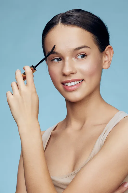 Young woman applying brow gel, showcasing beauty routine with a serene expression against a soft blue background, highlighting natural beauty and skincare trends