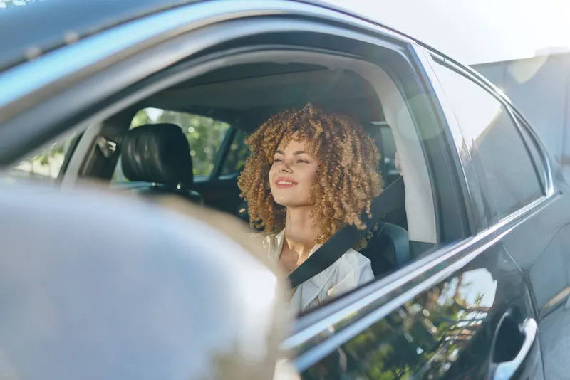 Smiling woman with curly hair wearing white blouse sitting in car seatbelt fastened, enjoying sunny day with relaxed expression and natural light inside vehicle.