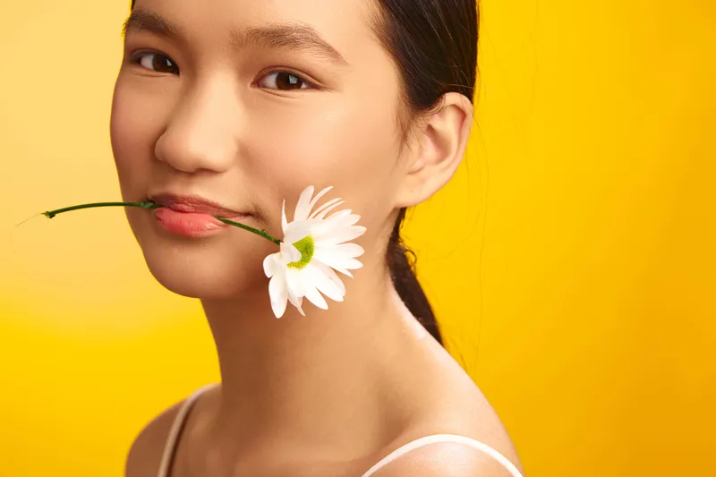 asian woman radiates a gentle smile as she holds a white flower near her lips against a bright yellow backdrop, creating a fresh, graceful, and contemporary mood.
