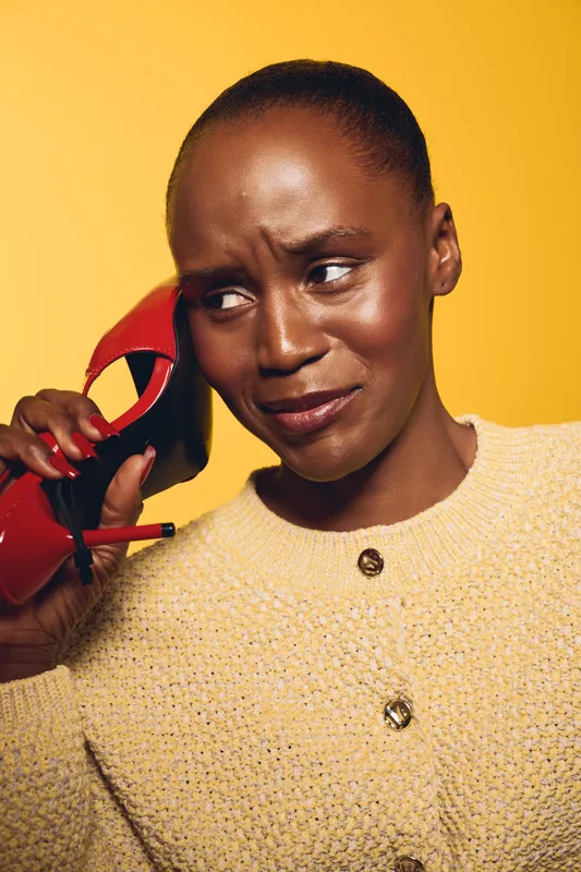 Portrait of a dark skinned African woman with short hair holding a red high heel shoe near her ear. She wears a beige sweater and looks confused against a yellow background.