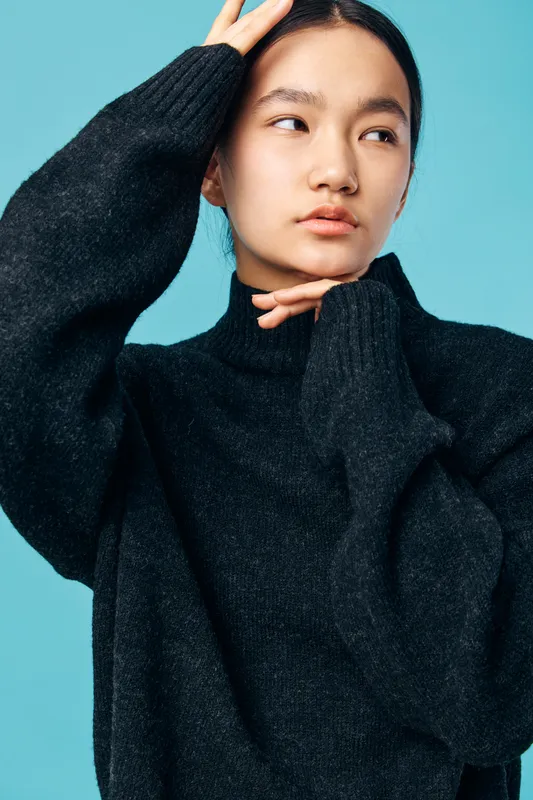Young woman in black sweater posing with hand on head and chin against blue background. Fashion, model, casual style, contemplative expression, studio photography in portrait format.