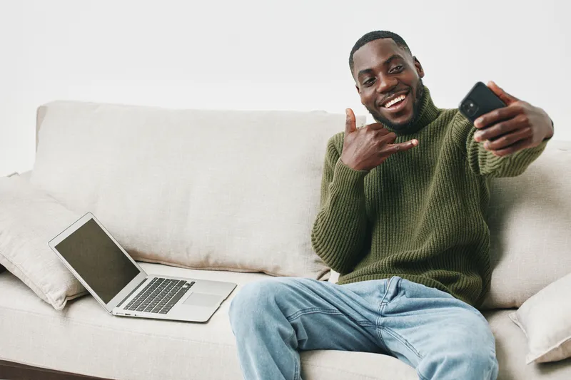Happy young Black man taking a selfie on the couch, wearing a green sweater, with a laptop nearby, showcasing a relaxed and joyful home environment