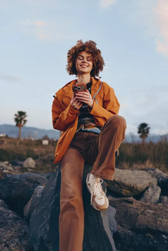Woman lifestyle outdoors, curly hair woman in orange jacket sits on rocks holding phone, smiling at sunset, relaxed posture, natural light, travel moment, blurred landscape background