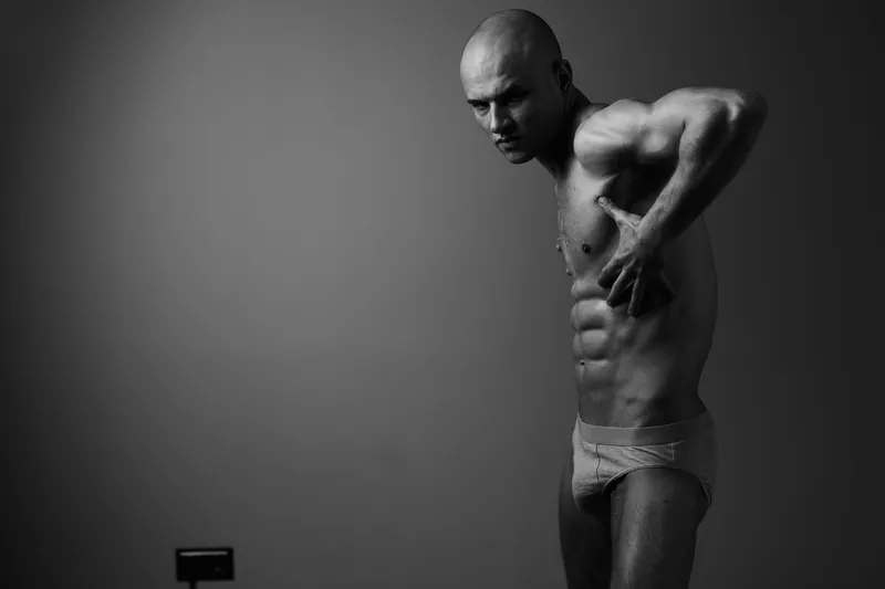 Black and white image of a muscular man presenting his strong physique in an artistic pose, emphasizing body definition and strength in a minimalist setting.