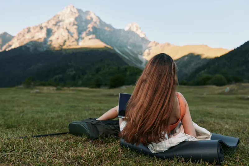 A person with long hair sits on a blanket in a grassy meadow with towering mountains in the background, working on a laptop and enjoying the outdoors.