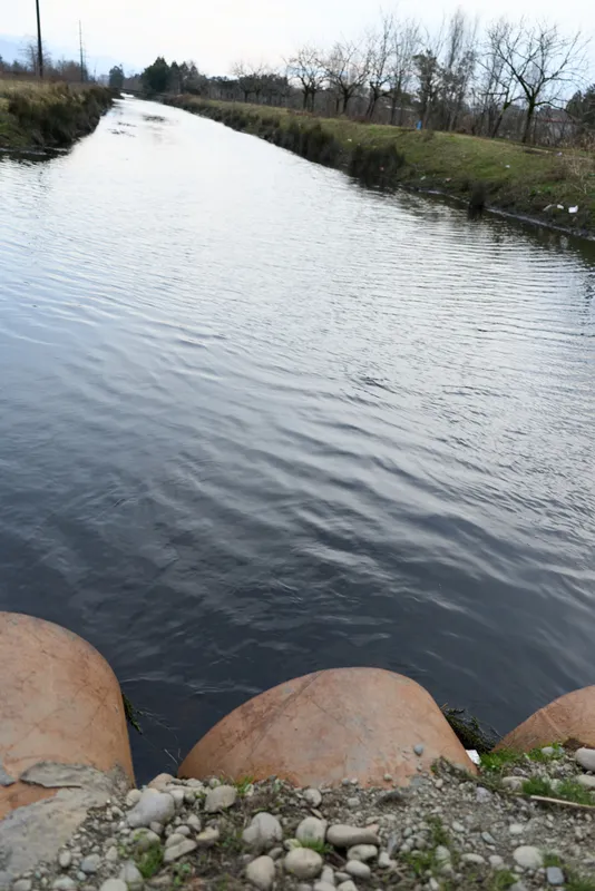 River with calm water and natural shoreline, a woman lower legs resting on stones at the bank. Outdoor landscape with leafless trees, cloudy daylight. Nature, relaxation, lifestyle.