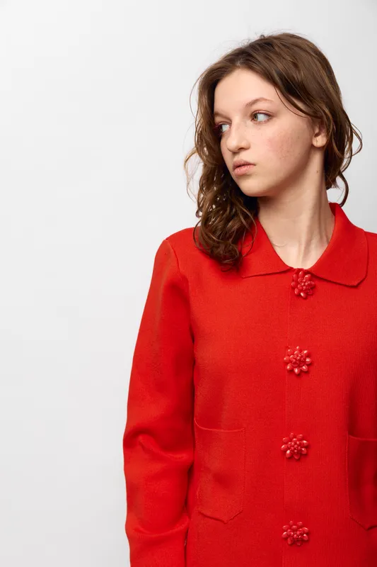 Young woman with curly hair wearing a red knitted sweater looks to the side with a neutral expression. Studio portrait on white background with soft lighting.