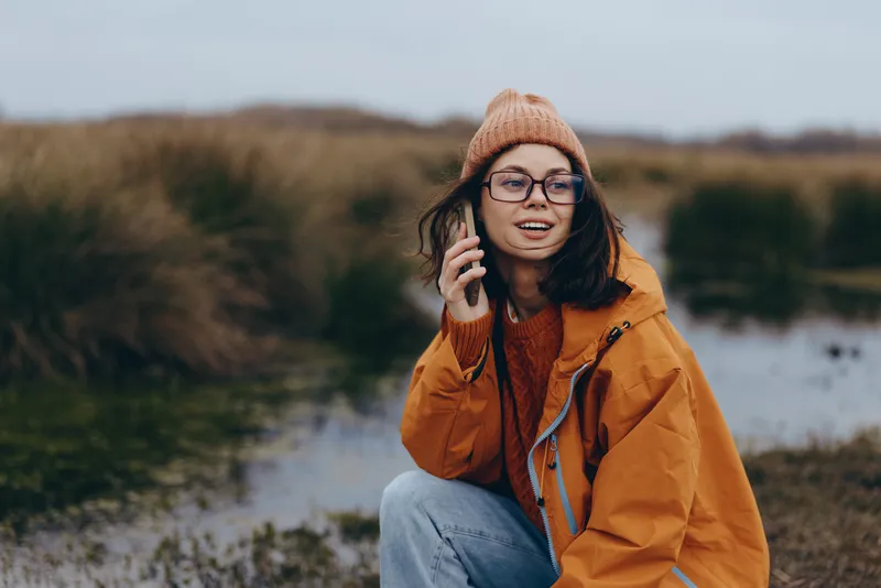 Woman lifestyle outdoors, smiling, phone held to ear, wearing orange jacket and knit hat, winter casual portrait by riverbank, calm moment with scenic hills and water