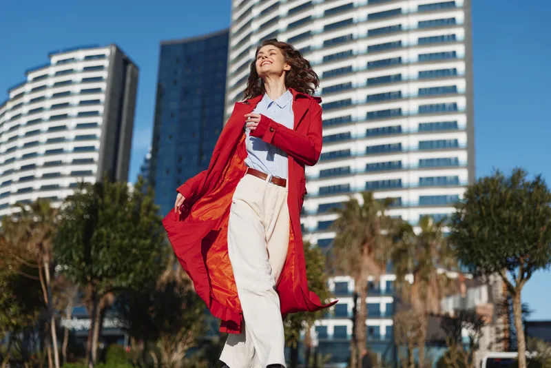 Cheerful woman outdoors wearing a bright red coat, confident pose, sunlit day with an urban skyline behind, stylish casual look, wind catching fabric