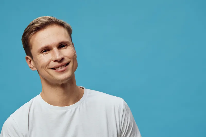 Smiling man with short brown hair wearing casual white t-shirt, looking at camera, standing against plain blue background, studio portrait, people lifestyle concept, positive expression, cheerful mood