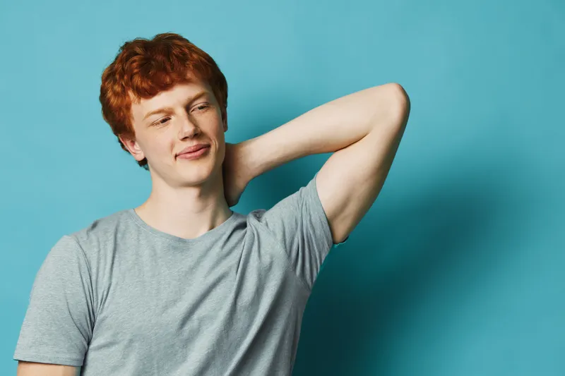 Redhead man wearing gray t shirt stands looking away with hand behind neck on blue background smiling and relaxed casual style portrait.
