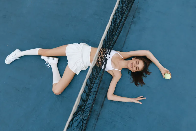 Playful young woman in white sportswear lying on a blue tennis court, smiling and holding a tennis ball, showcasing a vibrant and energetic atmosphere