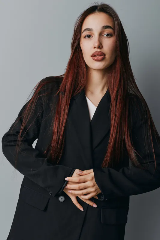 Young woman with long hair wearing black blazer posing with hands clasped in front of gray background. Portrait of confident female model with natural makeup and serious expression.
