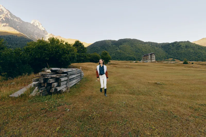 Woman walking across a wide meadow and open field with distant mountains in the background, nature landscape in a rural setting, casual outfit with boots and jacket for outdoor exploration.