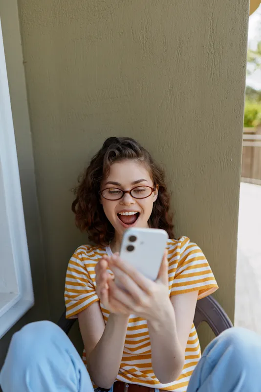 A young woman with curly hair and glasses sits on a sunny porch, laughing at her phone in a striped shirt, conveying candid everyday joy and relaxed home moments.