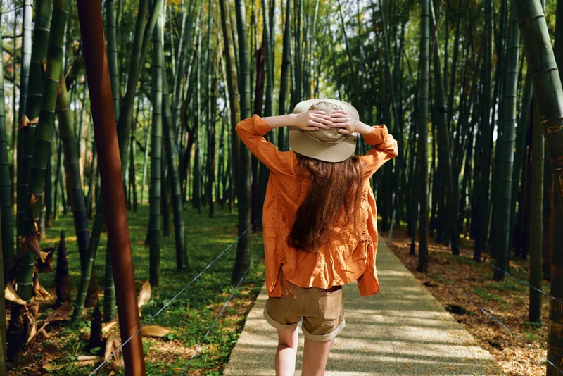 Woman walking on a path through a bamboo forest, wearing a hat and orange shirt, back view of a casual traveler enjoying nature and sunlight on a peaceful outdoor trail.