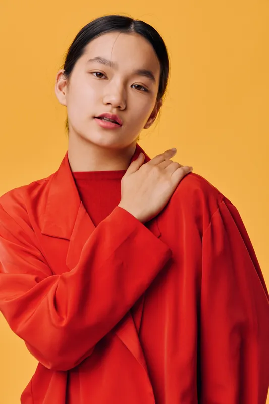 Young woman with black hair wearing red oversized jacket posing on orange background looking at camera with soft expression and hand on shoulder. Studio portrait with vibrant colors.