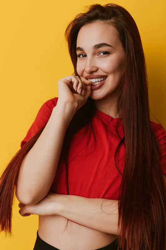 Young woman with long hair wearing red shirt smiling and posing with hand near mouth on yellow background. Portrait of happy female model with natural makeup and casual style.
