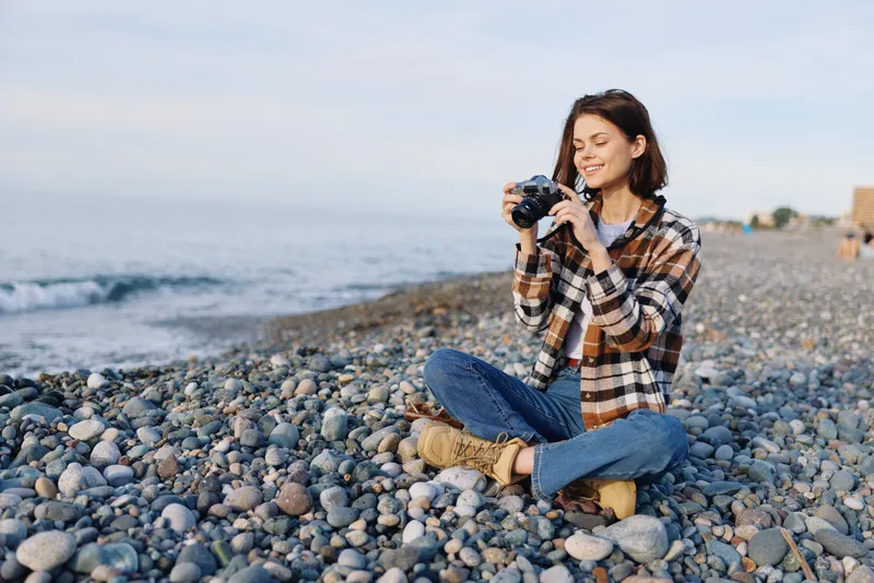 Female photographer crouches on a pebbled beach with a camera, wearing a plaid shirt and jeans, capturing the tranquil seaside scene and casual outdoor moments in daylight and breeze.