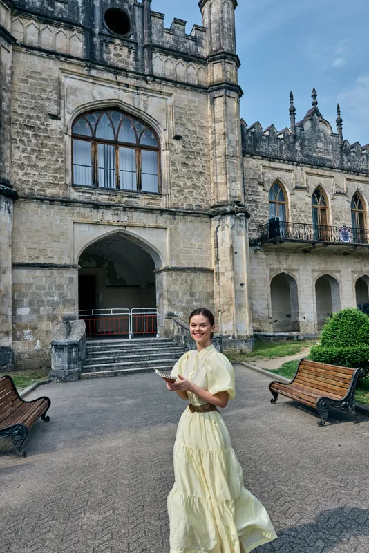 Woman in an elegant dress stands outdoors near a historic castle, holding a notebook in a sunlit courtyard, highlighting fashion, architecture, and timeless style in a serene setting.