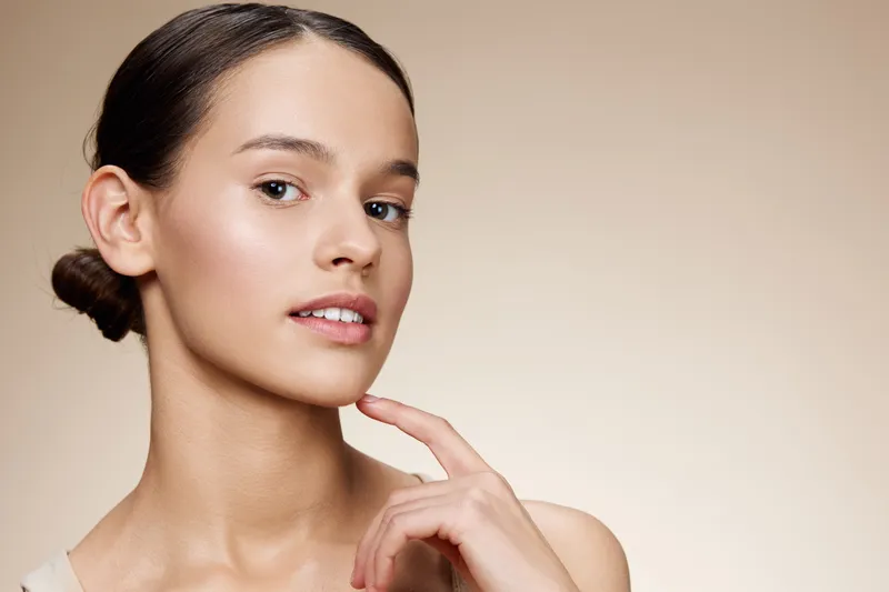 Natural beauty portrait of a young Caucasian woman with a soft expression, showcasing clear skin and minimal makeup against a warm beige background