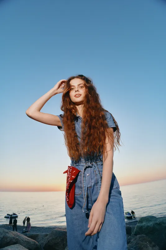 Lifestyle woman in modern boho-western denim street style stands confidently by the ocean at sunset, featuring natural red hair and relaxed film color tones.