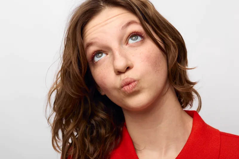 Young woman with brown hair in red shirt looks up with thoughtful expression on white background. Concept of curiosity, thinking, and contemplation in casual portrait.