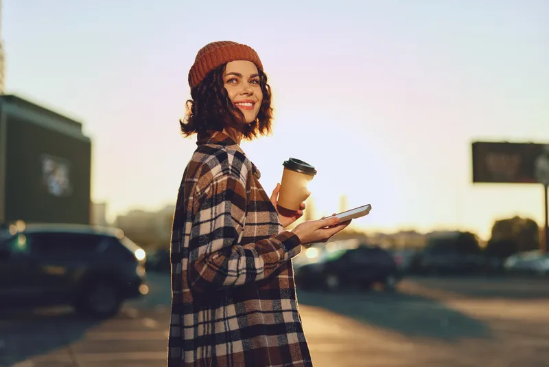 young woman, coffee cup, smartphone, city street. Authentic candid lifestyle portrait of a smiling woman in beanie at golden hour glow, mindful living and emotional storytelling.