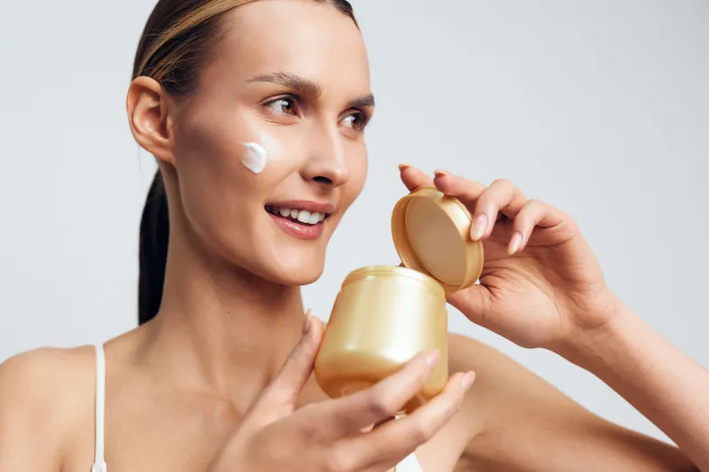 A smiling woman with clear skin applies face cream from a golden jar in a bright studio. The concept focuses on skincare, beauty routine, and healthy skin care products.
