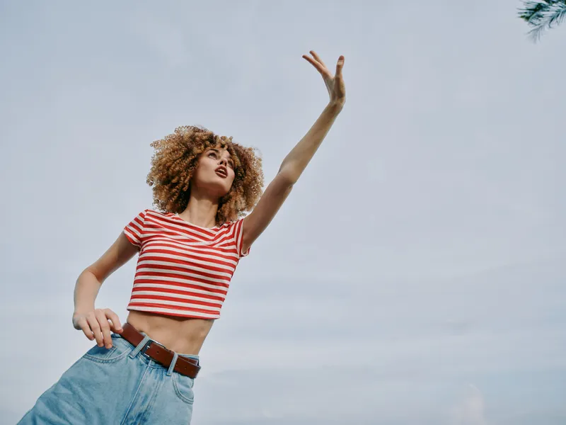 Female dancer outdoors with curly hair, wearing a striped crop top and denim jeans, reaching upward with outstretched arm, capturing energy, freedom, and springtime joy.